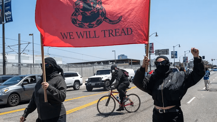 Gadsden flag variant
"We Will Tread" flag
Protest imagery
Political demonstration
Activist protest
Street protest
Black bloc tactics (implied by attire)
Anarchist symbolism (implied)
Anti-establishment protest
Urban protest scene
Political messaging
Social unrest imagery
Counter-protest (if context implies)
Symbolic flags
Contemporary protest movements
Disobedience (implied)
Resistance (implied)
Key Phrases:

"We Will Tread" protest flag
People demonstrating with Gadsden flag variation
Protestors in black attire
Political protest on city street
Symbolism of "We Will Tread"
Images of modern protests
Activism with alternative Gadsden flag
Protest against authority (implied)
Masked protestors with banners
Street demonstration with political messaging
Detailed Description for SEO:

This image captures a street protest scene featuring several individuals, some with faces obscured by black coverings or masks. Prominently displayed in the foreground is a large red flag featuring a coiled snake design above the text "WE WILL TREAD," a clear inversion of the traditional "Don't Tread on Me" Gadsden flag. This modified Gadsden flag, with its altered slogan, often signals a more assertive or confrontational stance, potentially associated with anti-establishment or anarchist-leaning movements. A person on a bicycle is also visible in the background, further indicating a public demonstration or march. The urban setting, with cars and buildings lining the street, provides context for the public nature of the protest. The attire and visual messaging strongly suggest an activist demonstration with a defiant political message.